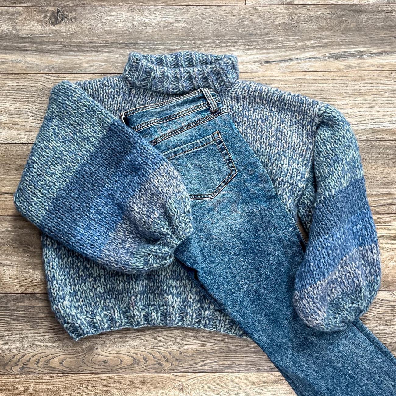 Denim blue melange sweater with a rolled collar, laid flat on a wooden floor beside a pair of jeans