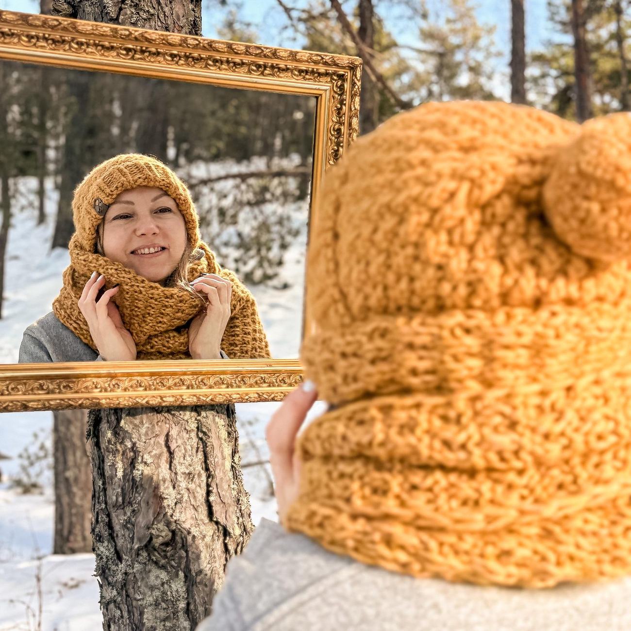 Agnes smiling in the snowy forest, wearing the honey yellow scarf and beanie, holding the gilded frame around herself