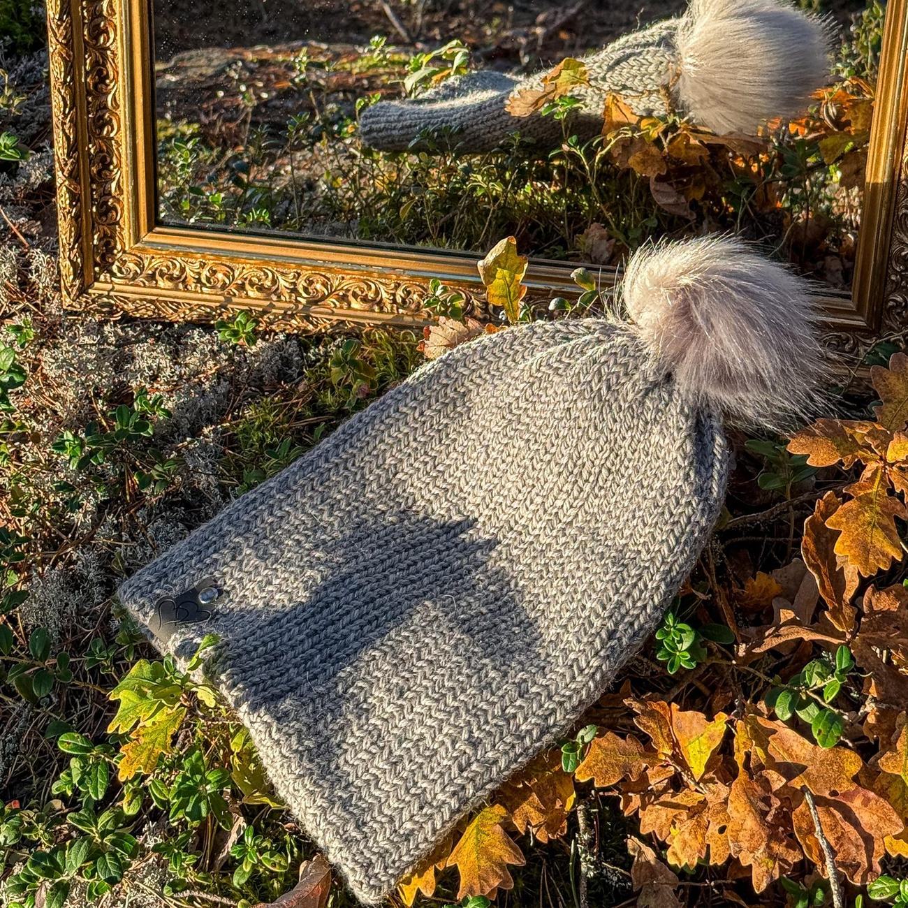 Light grey herringbone knit beanie with a big beige faux-fur pom, lying on moss and autumn leaves by the mirror
