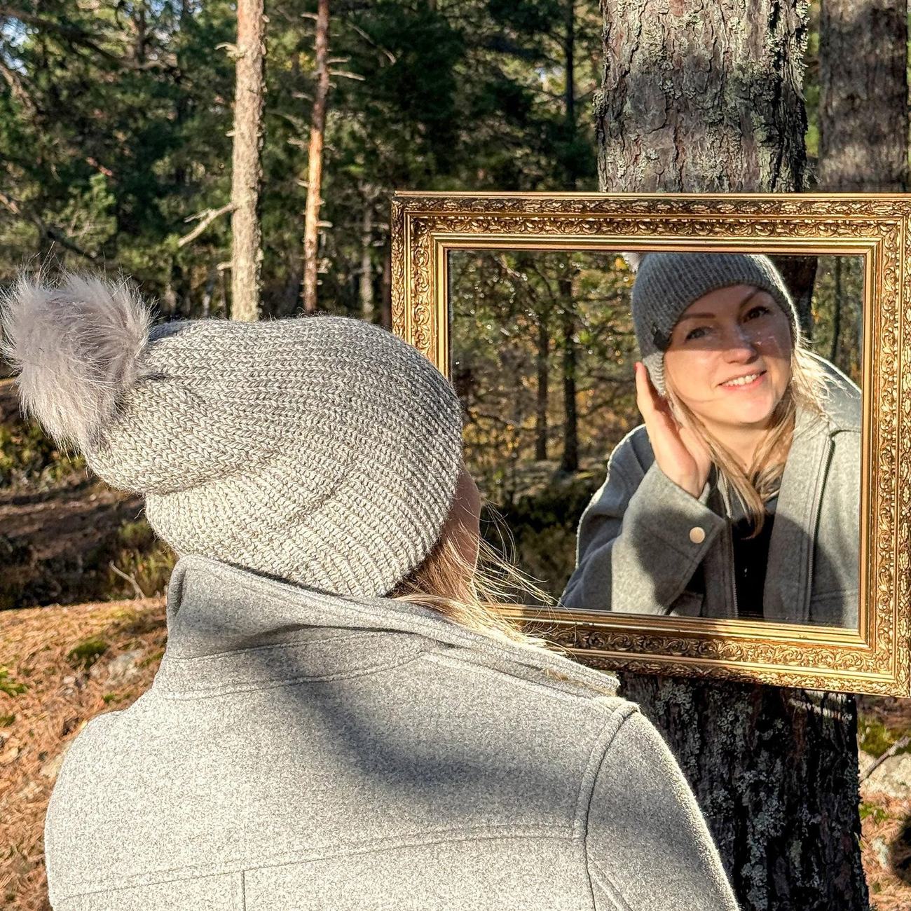 Agnes framed in the gilded mirror among the pines, wearing the grey beanie