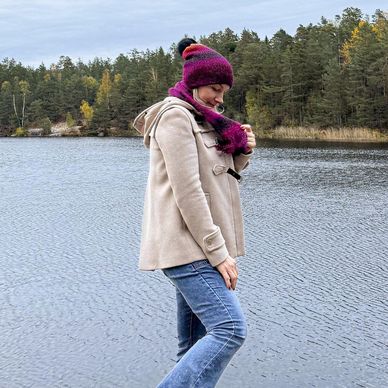 Walking along a Nordic lake in the hand-knitted Magenta to Midnight beanie and scarf, pine forest across the water