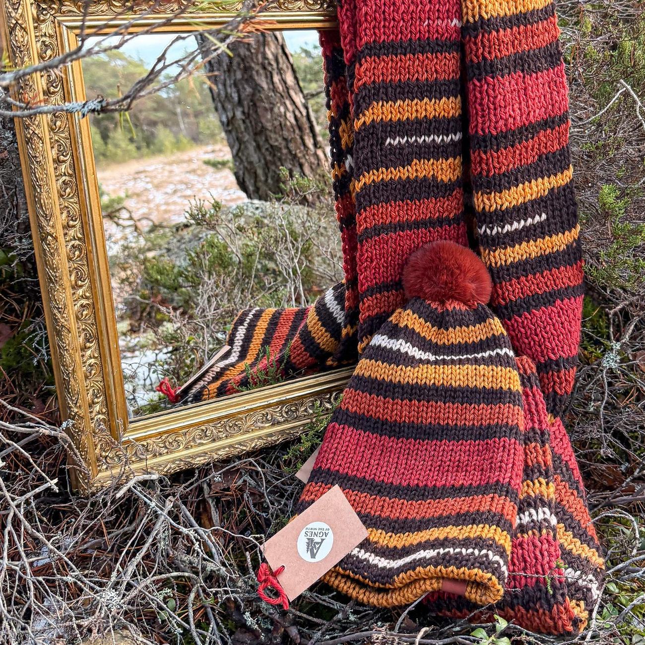 Beanie and scarf set in Autumn Stripes, brick red, ochre, chocolate and cream bands with a rust pom, draped over the gilded mirror in the woods